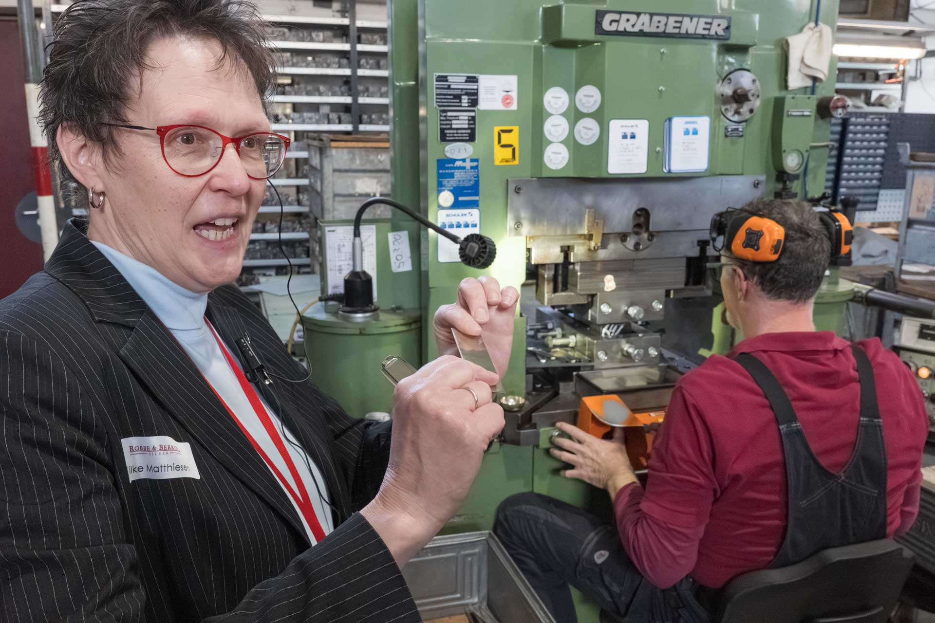 Eine Frau zeigt ein Stück Metall vor eine Werkbank